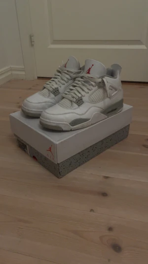 Nike Air Jordan 4 oreo - Nike Air Jordan 4 sneakers i vit och grå färg med röda detaljer på tungan. Skorna har snörning, meshpaneler på sidorna och en platt sula med stänkmönster. Ikonisk Jumpman-logga på hälen och tungan. Perfekt för dig som gillar klassiska och stilrena sneakers.