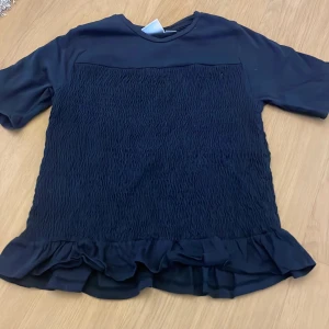 Mörkblå topp med volang från Gina Tricot - Säljer en mörkblå topp från Gina Tricot med korta ärmar och volangdetalj nedtill. Toppen har en smockad struktur framtill och baktill som ger en snygg passform. Perfekt att styla till jeans eller kjol! Haft i ett halvår men är endast använd några gånger. Skulle säga att den passar xs också! 