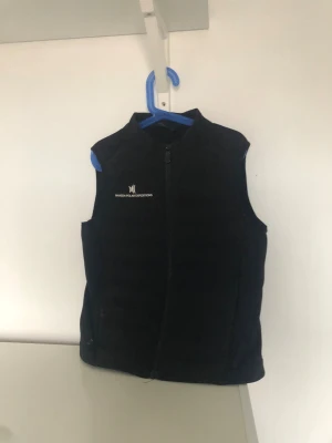 Svart dunväst från Nansen Polar Expeditions - Svart dunväst med hög krage och dragkedja framtill från Nansen Polar Expeditions. Västen har ett stilrent, quiltat mönster och en diskret logga på bröstet. Materialet är mjukt och lätt, perfekt för lager-på-lager. Snyggt präglad detalj på ryggen.