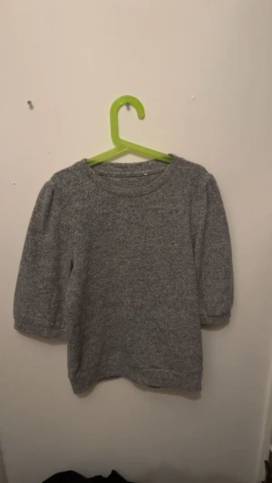 Grå stickad tröja med puffärm - Mysig grå stickad tröja med rund hals och puffade trekvartsärmar. Perfekt för dig som gillar enkel men snygg stil. Materialet är mjukt och värmande, och färgen är neutral vilket gör den lätt att matcha med andra plagg.