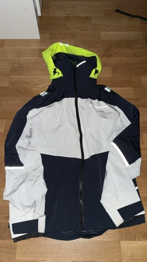 Ocean Works vindjacka med luva - Snygg vindjacka från Ocean Works i mörkblått och ljusgrått med detaljer i neon-gult. Jackan har hel dragkedja, reflekterande detaljer och justerbar huva. Perfekt för blåsiga dagar och outdoor-aktiviteter. Materialet är lätt och syntetiskt.