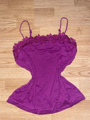 Lila linnetopp med spetsdetaljer - Säljer en lila linnetopp med tunna axelband och dekorativ spets längs överkanten. Toppen har en feminin och figurnära form, perfekt för att skapa en snygg look. Materialet känns mjukt och lätt mot huden.