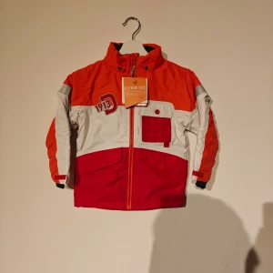 Didriksons vinterjacka - Snygg vinterjacka från Didriksons 1913 i rött, orange och vitt med coola reflexdetaljer och dragkedja framtill. Jackan har huva, fickor och är tillverkad i slitstark polyester med vattentätt Stormsystem™. Perfekt för kyliga och regniga dagar.