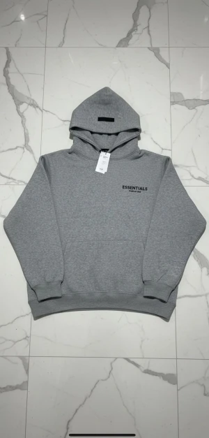 Grå Essentials Fear of God hoodie - Snygg grå hoodie från Essentials Fear of God med svart logga på bröstet och stor huva. Tröjan är i mjuk bomullsblandning, har ribbade muddar och klassisk oversized passform. Perfekt för en chill och stilren look.