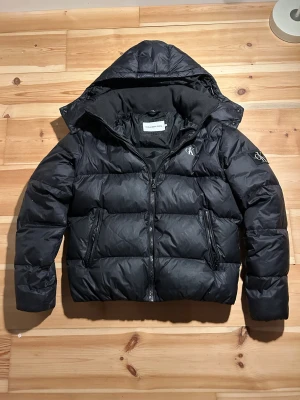 Calvin Klein pufferjacka - Fräsch och nästan oanvänd Calvin Klein pufferjacka som passar bra till vintern. Den är varm och har fodring kring halsen för extra värme. Luvan är även löstagbar. Skriv vid frågor, priset är inte hugget i sten :)