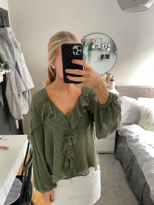 Olivgrön blus med volang och knyt - Säljer en olivgrön blus med v-ringning, volangdetaljer och knyt med tofs framtill. Blusen har diskret prickigt mönster, långa puffiga ärmar med resår vid handlederna och är tillverkad i ett lätt, luftigt tyg. Perfekt för dig som gillar romantiska och boho-vibbar.