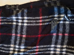 Rutig ullhalsduk från Burberrys - Snygg rutig halsduk från Burberrys i 100% lammull. Mörkblå botten med vita, röda och grå rutor. Klassisk design med fransar i ändarna, perfekt att slänga över axlarna för en cool och tidlös look.