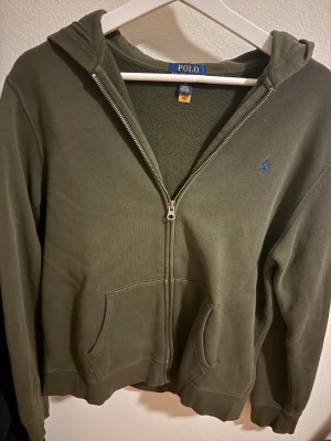 Olivgrön hoodie från Polo Ralph Lauren XL - Olivgrön hoodie från Polo Ralph Lauren i storlek XL. De är inte vanlig XL, skulle tror att de passar mellan 160-170cm skriv för mer information.Tröjan har dragkedja, huva och två fickor framtill. Broderad blå logga på bröstet. Tillverkad i mjuk bomull med fleece på insidan, perfekt för chill dagar. Extremt bra skicka och märks knappt att den är använd. Tveka inte på att höra av dig om pris eller information