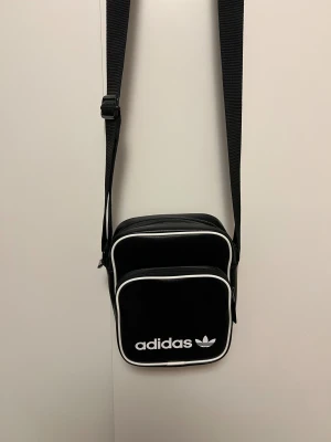 Svart Adidas axelväska - Svart  axelväska från Adidas med vit logga och text framtill. Väskan har två fack med dragkedja och vit kant runtom. Justerbar axelrem i svart och stilren design. Materialet är syntet med lätt struktur. Perfekt för att bära det viktigaste. Använde den ett få tal gånger. Ny kostade den 700kr.
