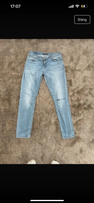 Ljusblå Nudie Jeans, storlek 29/30 - Säljer ett par ljusblå jeans från Nudie Jeans i rak modell. Klassisk femficksdesign med snygga slitningar och diskreta revor på framsidan. Jeansen har broderad detalj på bakfickan och är tillverkade i mjukt bomullsmaterial. Perfekt passform för dig som gillar en avslappnad stil.