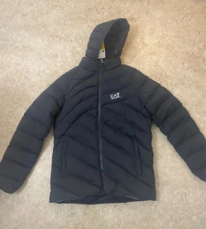 Svart pufferjacka från EA7 - Säljer en svart pufferjacka från EA7 med huva och snygga diagonala quiltade sömmar. Jackan har dragkedja framtill och två fickor på sidorna. Perfekt för dig som vill ha en stilren och varm jacka med sportig vibe.