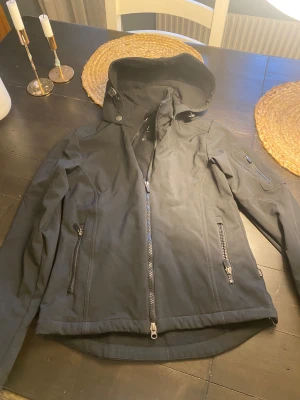 Svart vind/ridjacka  från CRW XS - Svart vind/rid jacka från CRW med luva och snygga dragkedjor fram och på fickorna. 