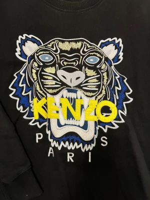 Svart Kenzo sweatshirt - Svart sweatshirt från Kenzo med ikonisk broderad tiger i blått, vitt och gult på bröstet. Tröjan har rund hals, ribbade muddar och lång ärm. Stort gult Kenzo-tryck och Paris-text under tigern. Perfekt statement-plagg för dig som gillar streetstyle.
