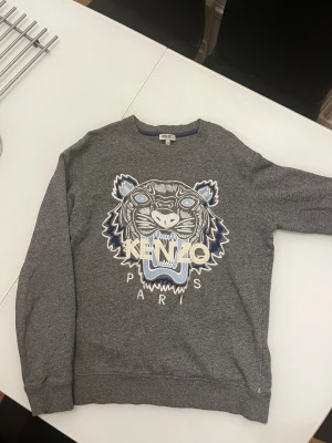 Grå sweatshirt från Kenzo - Säljer en grå sweatshirt från Kenzo med ikonisk broderad tiger i vitt och blått på bröstet. Tröjan har rund hals, ribbade muddar och lång ärm. Perfekt för dig som gillar statement-plagg och coola designer.