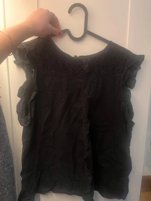 Svart blus med spets och volang - Säljer en svart blus med snygga spetsdetaljer upptill och volanger längs ärmarna. Blusen är ärmlös och har en rund halsringning. 