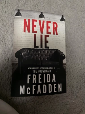 Never Lie - En spännande bok av Freida McFadden, författaren bakom succén The Housemaid. Omslaget med en mystisk soffa och dramatiska färger lockar till läsning för dig som gillar nervkittlande berättelser och oväntade vändningar.