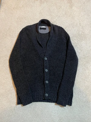 Mörkgrå stickad kofta med knappar | M - Snygg mörkgrå stickad kofta i grovt mönster med bred krage och fyra knappar framtill. Långärmad och ribbad vid ärmslut och nederkant. Perfekt för lager-på-lager och chill stil. Passar dig som gillar klassiska och enkla plagg.