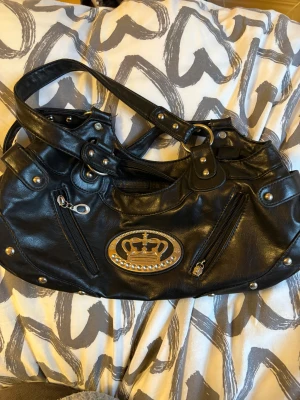 Svart handväska med kron-detalj - Cool svart handväska i skinnimitation med silvriga nitar och dragkedjor. Väskan har dubbla handtag och flera fack med dragkedja, perfekt för att hålla ordning på dina grejer.