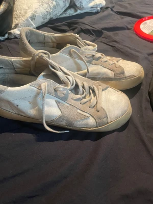 Golden goose med grå detaljer och stjärna - Säljer ett par vita sneakers med grå detaljer och en stor stjärna på sidan. Skorna har snörning och platt sula, ovandel i skinn och mocka. Klassisk streetstil med slitna vibbar och retrolook. Perfekta för dig som gillar en avslappnad och urban stil.