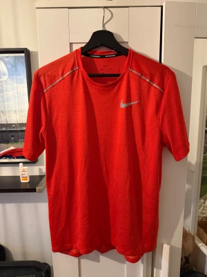 Röd Nike Dri-Fit t-shirt - Nike Dri-Fit t-shirt i stark röd färg med silverfärgad swoosh på bröstet och diskreta reflekterande detaljer vid axlarna. T-shirten har rund hals och är tillverkad i lätt polyester som andas, perfekt för löpning eller gym. Snygg och sportig look.