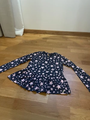 Mörkblå stjärnklänning från Ellos - Supersöt mörkblå klänning från Ellos med ljusrosa stjärnmönster över hela plagget. Klänningen har lång ärm, rund hals och en volang nertill som ger en lekfull look. Perfekt för dig som gillar stjärnor och vill ha något bekvämt och snyggt.