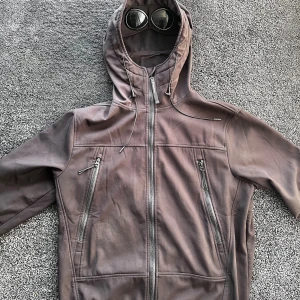 C.P. Company jacka - Säljer en brun Goggle Jacket från C.P. Company med ikoniska glasögon i huvan. Jackan har dragkedja framtill, två snedställda bröstfickor med dragkedja och justerbar huva med snörning. Ser brun ut på bilderna men är svart, Storlek L, skick 9/10, qr koden ör avklippt av förra ägaren så kan ej garantera äktahet därav priset. Pris går att diskutera✅