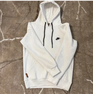 Vit hoodie från Nike med svart logo - Vit hoodie från Nike med svart broderad logga på bröstet och svarta snören i huvan. Tröjan har en klassisk känguruficka framtill och ribbade muddar vid ärmslut och nederkant. Perfekt för chill dagar eller när du vill ha en clean streetstyle.
