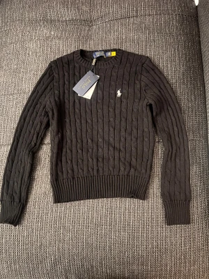 Svart kabelstickad tröja Polo Ralph Lauren - Svart kabelstickad tröja från Polo Ralph Lauren med klassisk rund hals och broderad logga på bröstet. Tröjan har långa ärmar och ribbade muddar vid ärmslut och nederkant. Perfekt för dig som gillar stilrena och tidlösa plagg.