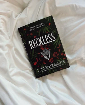 Reckless - En bok med en mörk och mystisk omslagsdesign, fylld med röda blomblad och gröna blad. Texten 'Traitor. Tormented. Destined for each other.' antyder intensiva känslor och dramatiska relationer. Perfekt för dig som gillar spännande och känsloladdade berättelser.