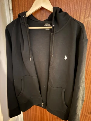 Svart hoodie från Polo Ralph Lauren - Svart hoodie från Polo Ralph Lauren med dragkedja, huva och snörning. Knappt använd. Har kvitto. Köpt för 2195kr