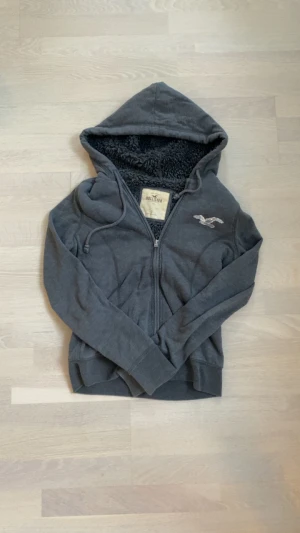 Grå fodrad hollister hoodie - Mysig grå teddyjacka från Hollister med huva och fluffigt foder. Jackan har dragkedja framtill, snörning vid huvan och ett silvrigt paljettmärke på bröstet. Perfekt för kalla dagar och chill outfits.