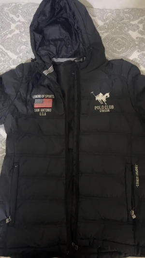 Svart pufferjacka från Vinson Polo Club - Svart pufferjacka från Vinson Polo Club med huva och broderade detaljer på bröstet. Jackan har dragkedja framtill, sidofickor med dragkedja och coola tryck som 'Legend of Sports' och 'Trophy Series'. Perfekt för kalla dagar och har en sportig vibe.