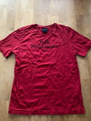 Röd Peak Performance t-shirt - Snygg röd t-shirt från Peak Performance med svart tryck på bröstet. Klassisk rund hals och korta ärmar, tillverkad i mjuk bomull. Perfekt för dig som gillar stilrena och sportiga plagg med tydlig logga framtill.
