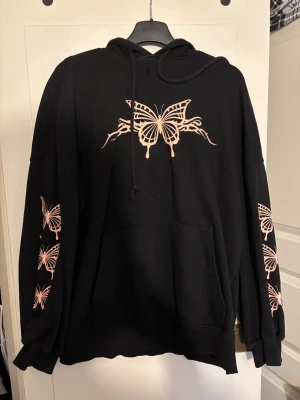 Svart hoodie med rosa fjärilar Divided - Svart oversized hoodie från H&M Divided med stora rosa fjärilar tryckta på bröstet och längs båda ärmarna. Tröjan har huva, snörning och en stor magficka. Mjuk insida och avslappnad passform, perfekt för chill dagar.