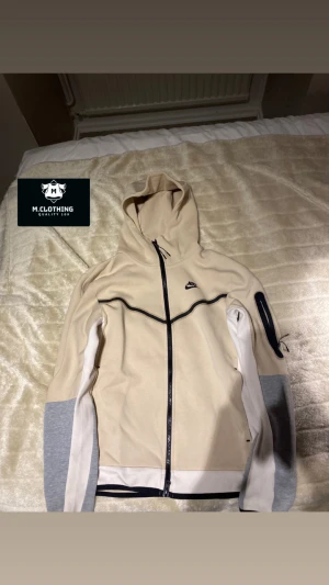 Beige Nike teck Old model  - Snygg beige hoodiejacka från Nike med svarta detaljer och hel dragkedja. Jackan har huva, gråa och vita partier på ärmarna samt en praktisk ficka med dragkedja på ena ärmen. Perfekt för en sportig och avslappnad stil. Passar M och S!