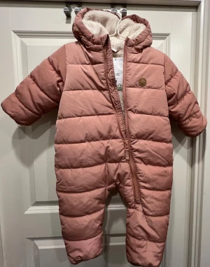 Rosa pufferoverall med öron och teddyfoder - Supermysig rosa pufferoverall från H&M med fluffigt teddyfoder och huva med söta öron. Overallens material är polyester och den har en sned dragkedja framtill för enkel påklädning. Perfekt för kalla vinterdagar och håller värmen ordentligt.
