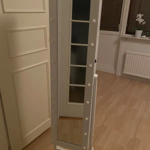 Smyckesskåp med spegel och LED-belysning - Stilrent smyckesskåp med helkroppsspegel och inbyggd LED-belysning. Skåpet har gott om krokar och hyllor för smycken och accessoarer. Vit finish, står stadigt på ben. Mycket gott skick, inga synliga defekter.Spegel med smyckesförvaring och den har ljus som man kan sätta på och den hämtas antingen hem till mig eller så betalar man bara varan via appen Tiptapp och sen så skickar jag den till dig via leverans skriv till mig så löser mig leveransen
