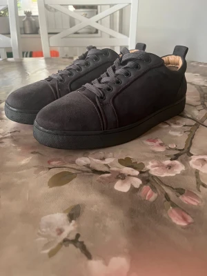 Svarta sneakers från Christian Louboutin - Säljer ett par svarta sneakers från Christian Louboutin med klassisk röd sula och rund tå. Ovandelen är i mjuk mocka med snörning och insidan har en beige skinninnersula. Snygg och stilren design som sticker ut med den ikoniska röda undersidan.