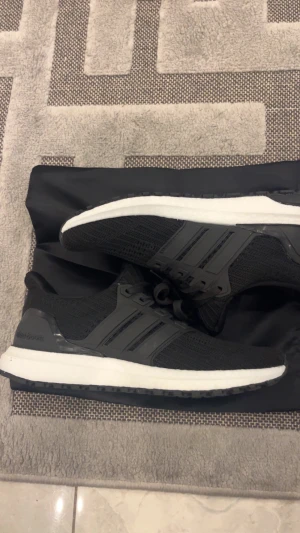 Svarta Adidas Ultraboost sneakers - Adidas Ultraboost sneakers i svart med vit platt sula. Skorna har stickad ovandel i syntetmaterial, klassiska tre ränder på sidan och snörning. Perfekta för dig som gillar stilrena och sportiga sneakers med grym komfort. Storlek 41