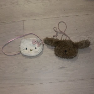 Axelväskor i mjuk plysch, barn  - Två gulliga axelväskor i mjuk plysch, en vit Hello Kitty med rosa axelrem och rosett, samt en brun väska formad som en hund med långa öron. Perfekt för barn som gillar söta accessoarer med djurtema.