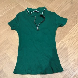 Grön ribbad polotopp från FB Sister - Säljer en grön ribbad polotopp från FB Sister i storlek S. Toppen har en cool dragkedja med ringdetalj framtill och en krage med vita och svarta ränder. Kortärmad och figurnära passform, perfekt för dig som gillar sportig och trendig stil.