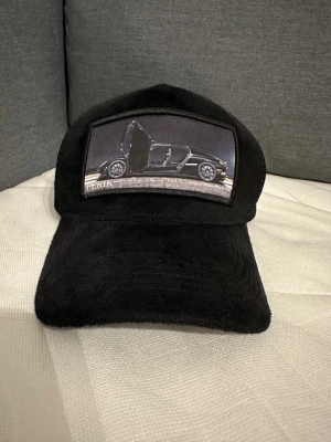 Svart keps med bilmotiv - Svart keps med mesh på sidorna och baksidan för extra ventilation. Framtill finns en patch med ett coolt bilmotiv där dörrarna är öppna. Materialet framtill är mjukt och har en sammetsliknande känsla. Perfekt för dig som gillar bilar och streetstyle.