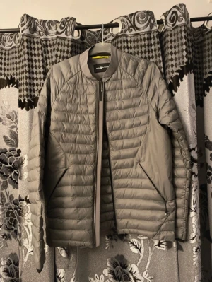 Grå pufferjacka från Sail Racing - Snygg grå pufferjacka från Sail Racing med stilren design och horisontella quiltade paneler. Jackan är i storlek M