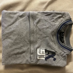 Armani Exchange Grå långärmad tröja - Snygg grå långärmad tröja från Armani Exchange med tryckt logga på bröstet. Tröjan har ribbade muddar och krage med blå och svarta ränder, samt en diskret bröstficka. Perfekt för dig som gillar stilrena och sportiga plagg.