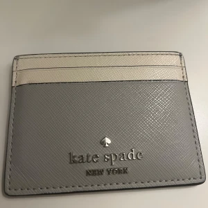 Kate Spade korthållare - Supersnygg kortplånbok från Kate Spade i grått och beige skinn. Stilren design med silverfärgad logotyp framtill och plats för flera kort. Perfekt att ha med sig i fickan eller väskan när du vill hålla det enkelt och classy. Äkta.
