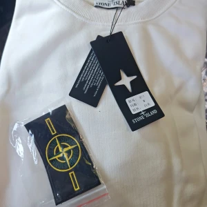 Vit sweatshirt från Stone Island - Vit sweatshirt från Stone Island med klassisk rund hals och ribbade muddar. Medföljer den ikoniska Stone Island-patchen som kan fästas på ärmen. Tröjan har en clean look och är perfekt för dig som gillar streetwear och snygga detaljer.  Dm vid intresse 