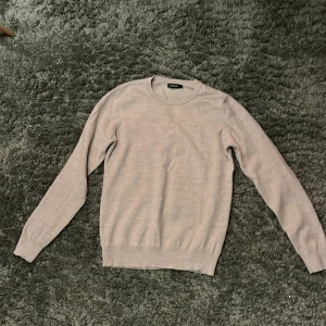 Beige stickad tröja från J.Lindeberg - Snygg beige stickad tröja från J.Lindeberg med rund hals och diskret logga på bröstet. Tröjan har långärmade ärmar och ribbade muddar vid ärmslut och nederkant. Perfekt för dig som gillar stilrena och klassiska plagg.