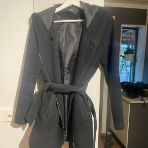 Svart kappa med huva och bälte - Svart kappa från John Boss med huva, knytskärp i midjan och dragkedja. Jackan har slät yta, fodrad insida och långa ärmar. Perfekt för dig som gillar stilren och enkel design med en touch av edge.