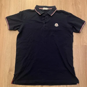 Mörkblå Moncler pikétröja - Snygg mörkblå pikétröja från Moncler med klassisk krage och korta ärmar. Tröjan har vita och röda detaljer på krage och ärmslut samt Moncler-logga på bröstet. Replika. Tillverkad i mjuk bomull för skön känsla. Perfekt för dig som gillar stilrena plagg med exklusiv touch.