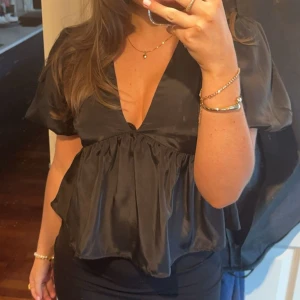 Svart blus med v-ringning - Säljer en svart blus i glansigt material med djup v-ringning och korta puffärmar. Blusen har en flowy, volangliknande nederdel som ger en snygg siluett. Från Nelly i storlek 38🩷